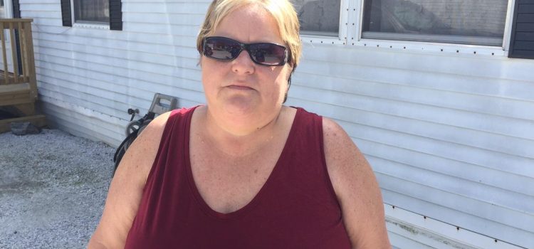 Blonde woman wearing sunglasses and a burgundy shirt stands in front of a mobile home.