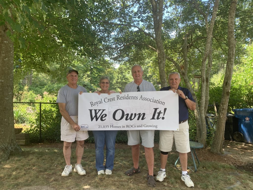 Four Royal Crest board members in front of tall trees in their community hold up a banner that reads "We Own It!"